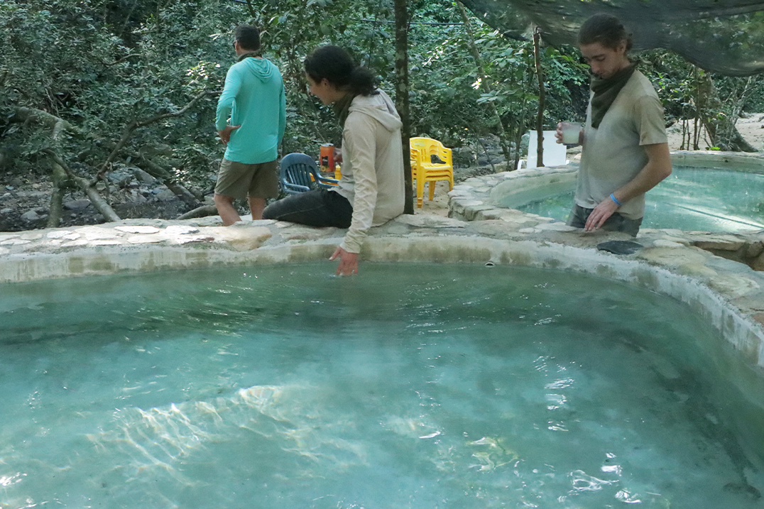 Personas relajándose en aguas termales naturales rodeadas de montañas y vegetación, disfrutando de una experiencia de bienestar en la naturaleza.