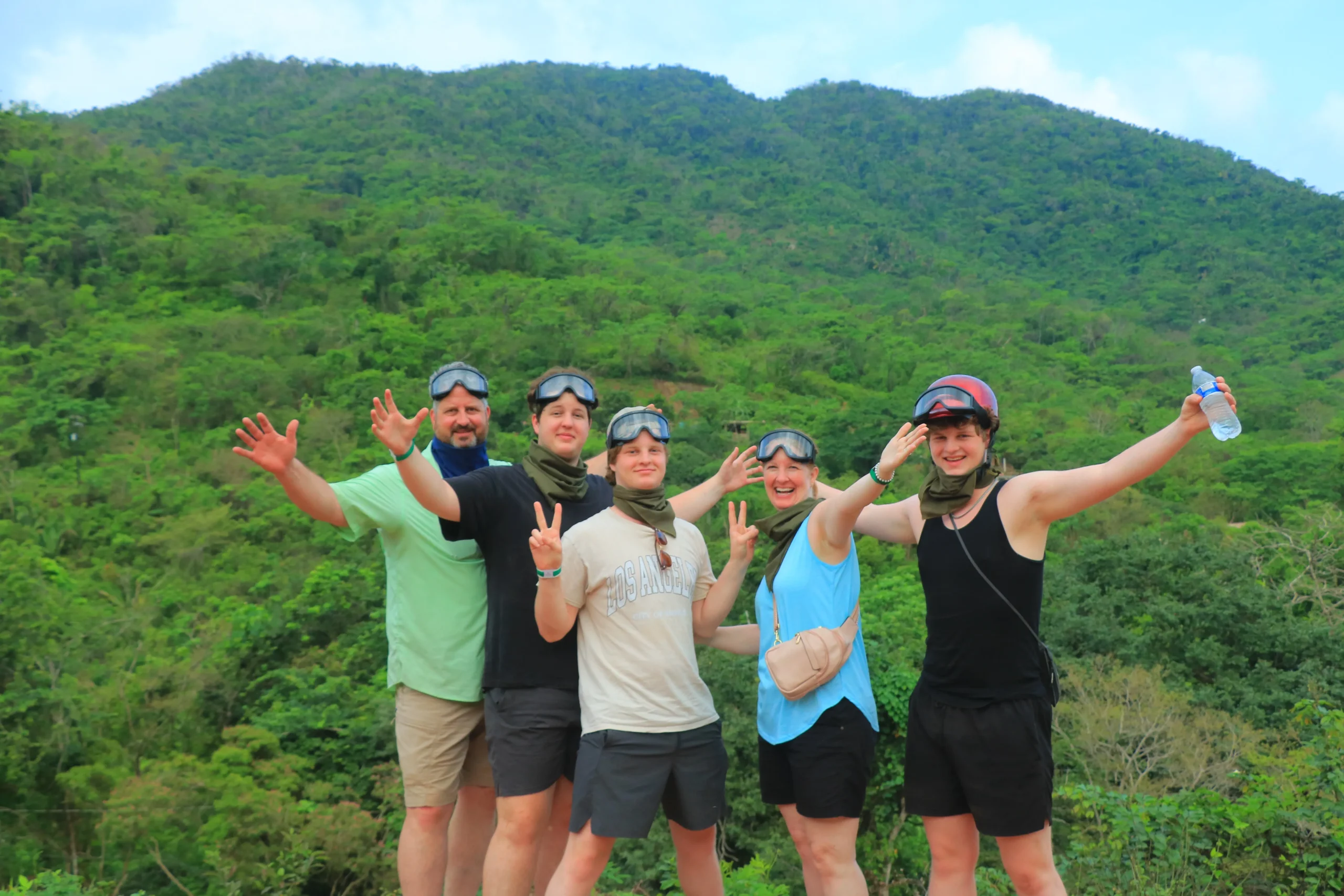 Five people who went on a RZR route pose with sports goggles in front of a mountain covered in greenery, enjoying the moment.