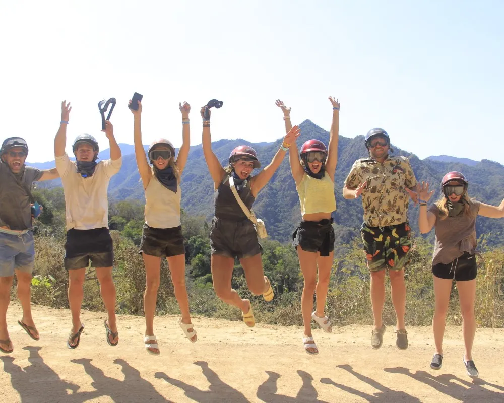 A group of people wearing helmets jumps with excitement on an all terrain trail surrounded by mountains.
