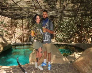 A cheerful moment of a couple holding drinks in a natural setting, with turquoise water in the background that conveys the atmosphere of Where to stay in Puerto Vallarta.