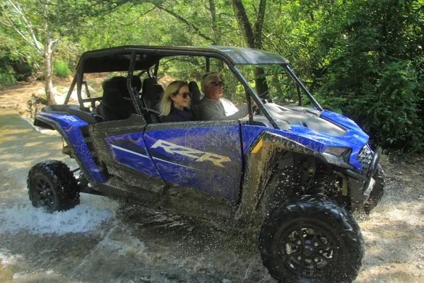 A group enjoys an adventure in a UTV, crossing a water-filled trail surrounded by tropical vegetation.